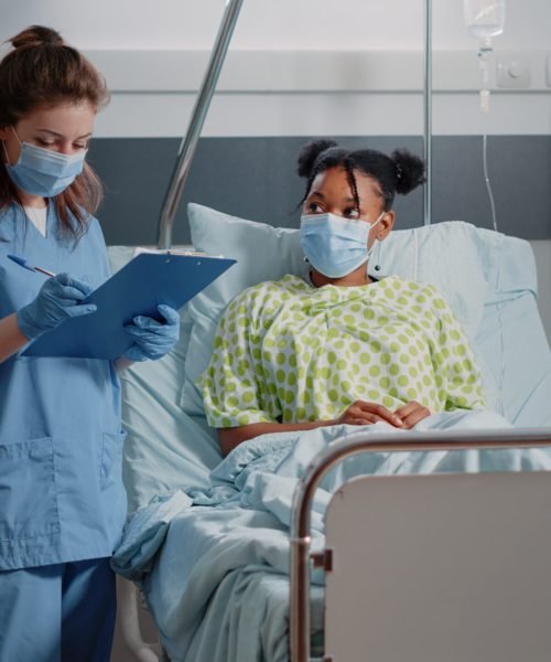 Medical assistant doing consultation with ill patient, wearing face masks in hospital ward. Nurse discussing with young woman about healthcare treatment and taking notes during pandemic